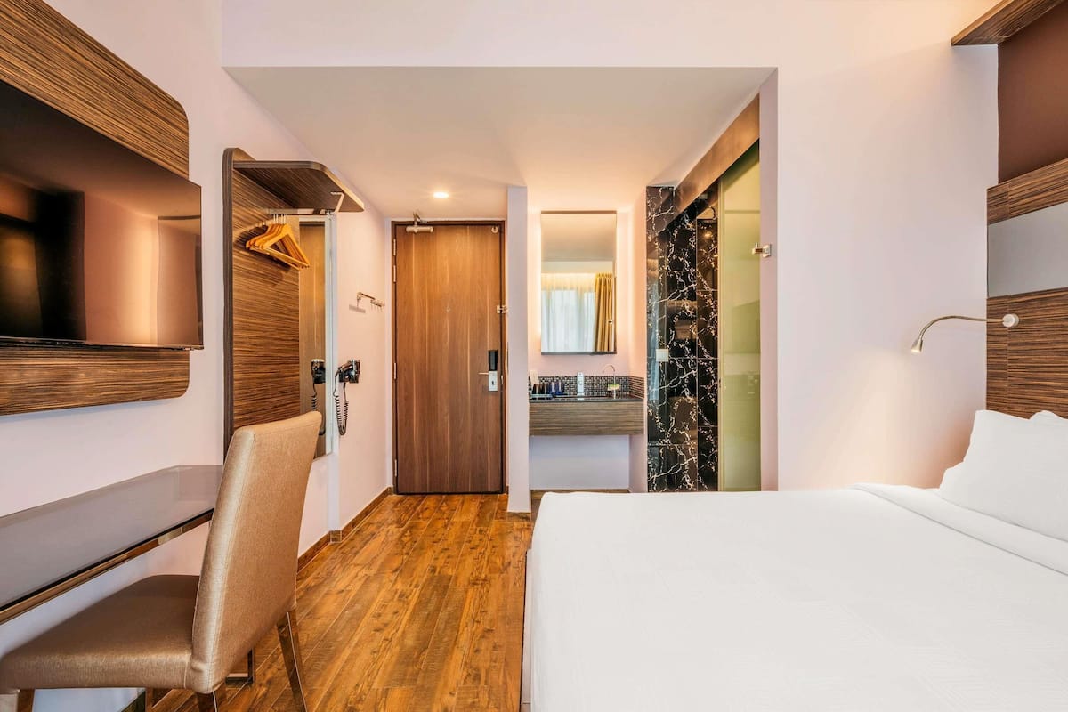 Habitación de hotel con cama, escritorio, silla, televisión, espejo y baño con ducha, además de piso de madera.