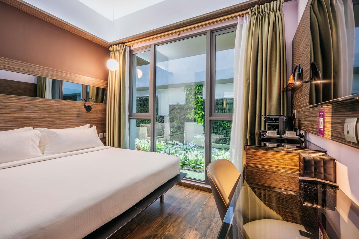 Habitación de hotel con una cama grande, un escritorio con cafetera y vistas a zonas verdes por las ventanas.