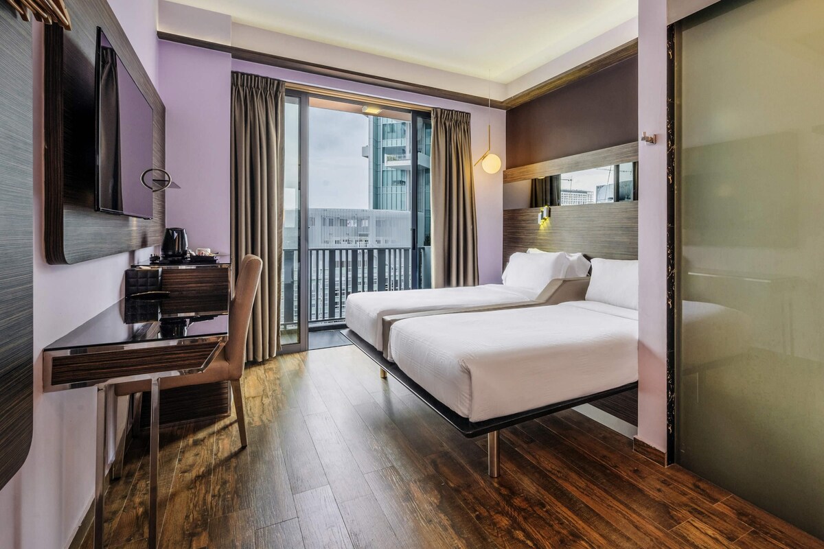 Habitación de hotel con cama, escritorio, silla, televisor y balcón con cortinas.