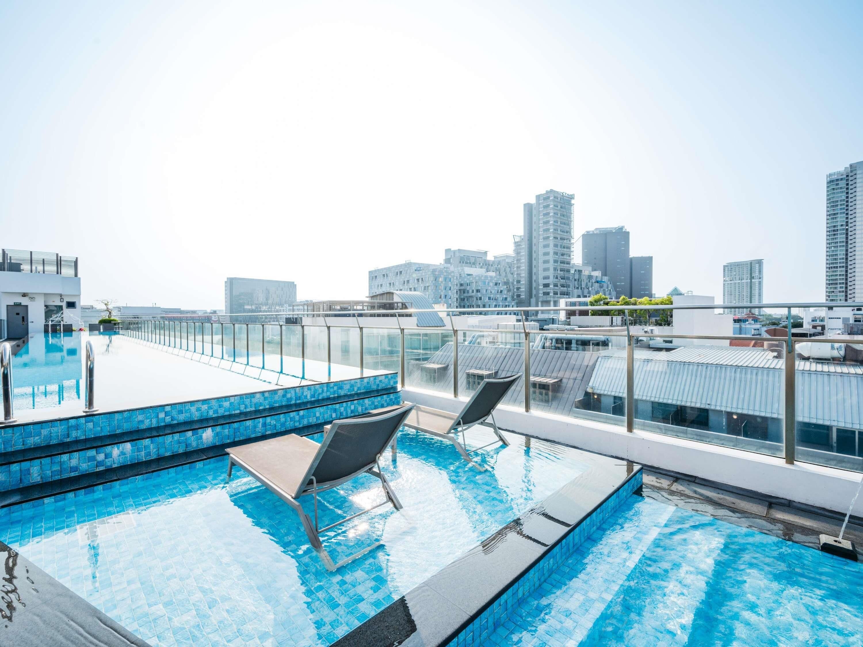 A rooftop infinity pool with a glass barrier, two lounge chairs, and a city skyline in the background.
