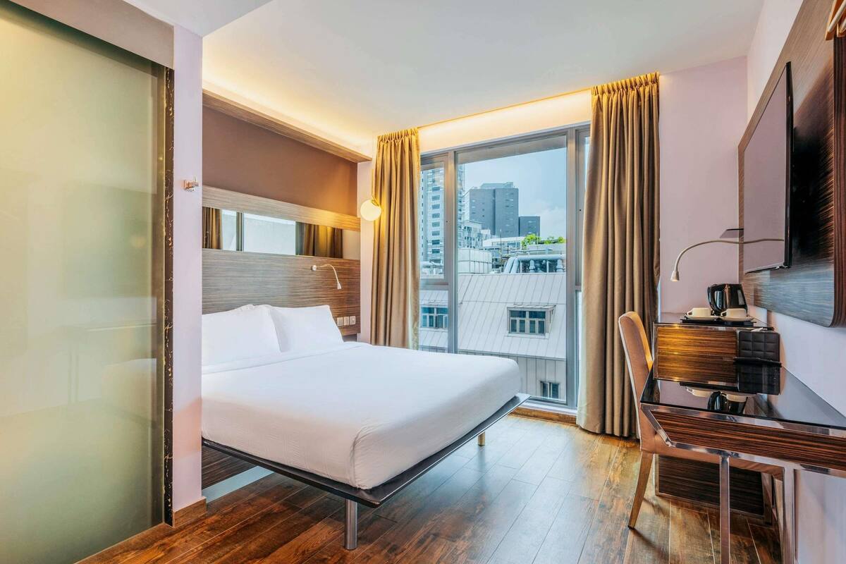 Una habitación de hotel con cama, escritorio, silla, televisor y un ventanal con cortinas.