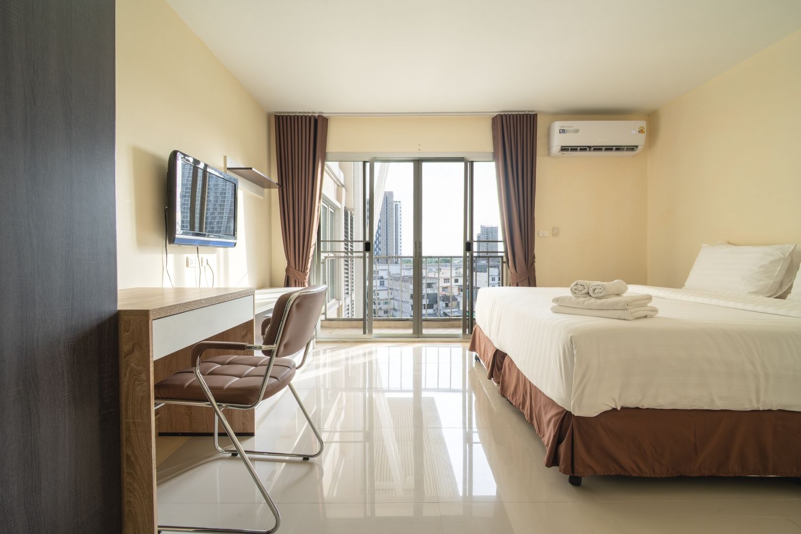 A hotel room with a bed, a desk, a chair, a television, and a view of the city through large windows.