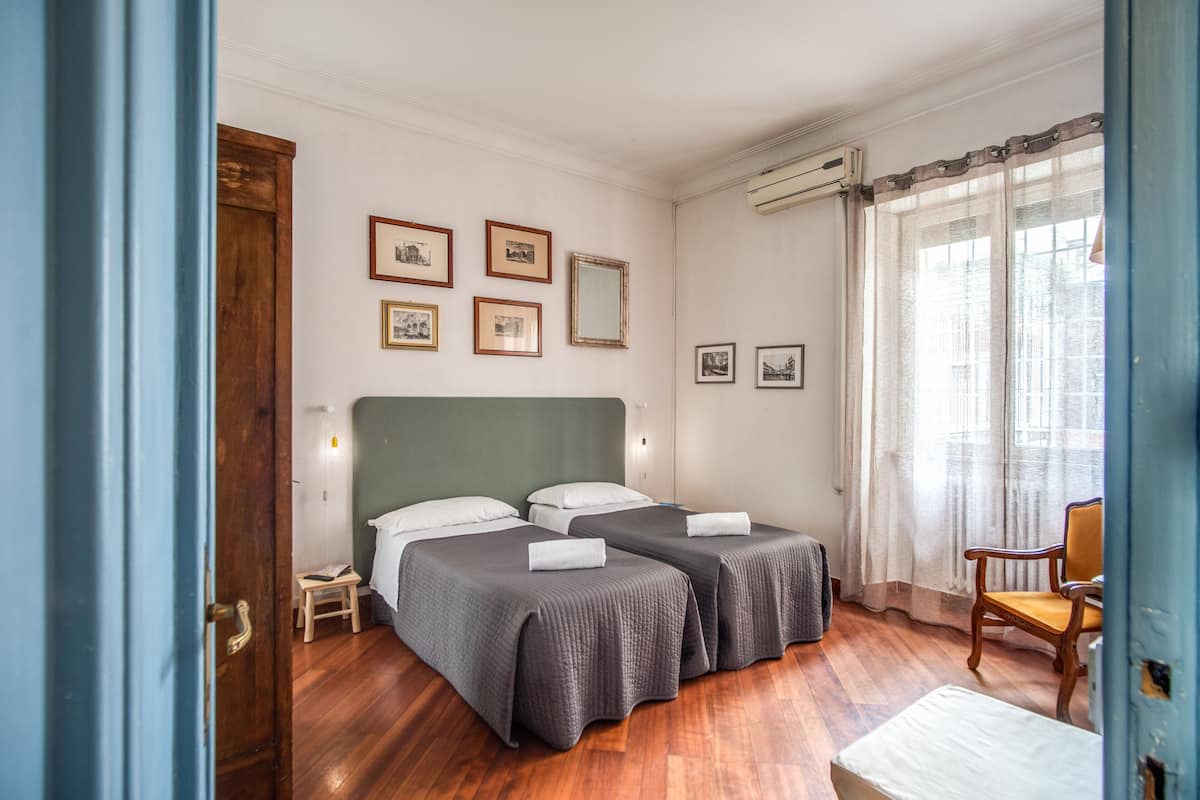 Una habitación con dos camas, suelo de madera, una silla y cuadros enmarcados en la pared.