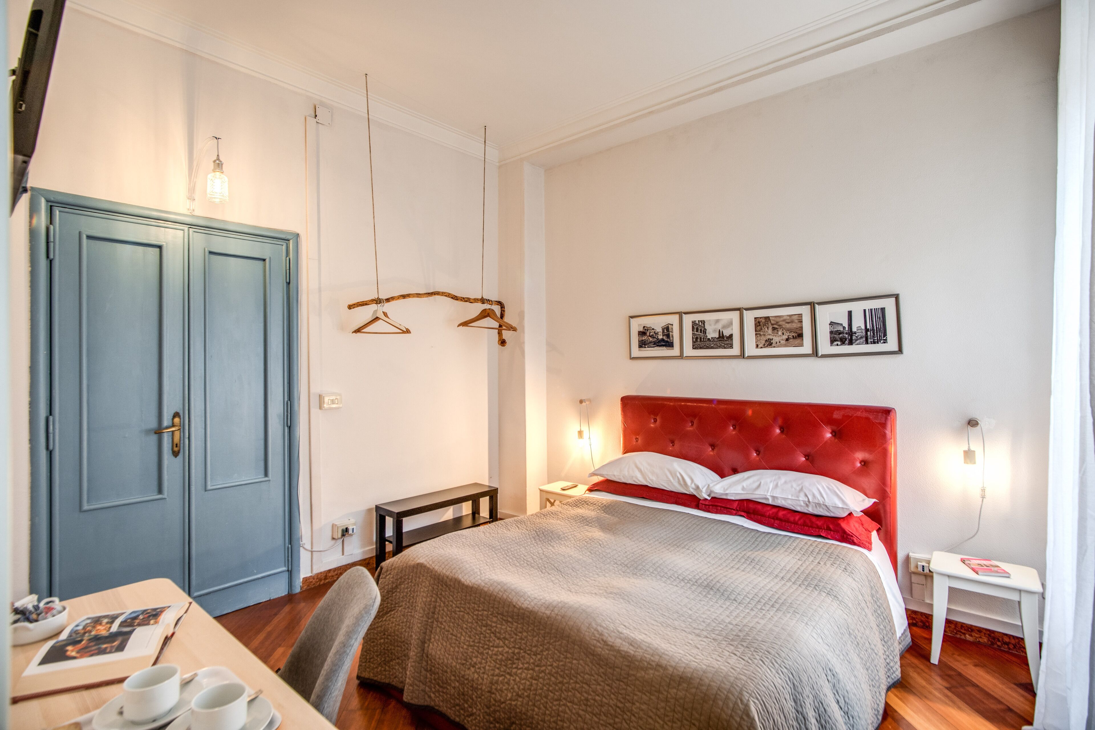Un dormitorio con cabecera roja, una cama con colcha gris y blanca, un escritorio de madera con una silla, un armario azul y una lámpara fijada a la pared.