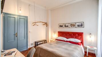 Un dormitorio con cabecera roja, una cama con colcha gris y blanca, un escritorio de madera con una silla, un armario azul y una lámpara fijada a la pared.