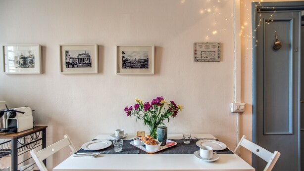 Un coin repas avec une table dressée pour deux personnes, des tableaux encadrés au mur et un vase rempli de fleurs.