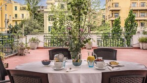 Un área de comedor al aire libre con una mesa dispuesta para dos personas, rodeada de sillas. Hay una planta en maceta con flores en el centro de la mesa.