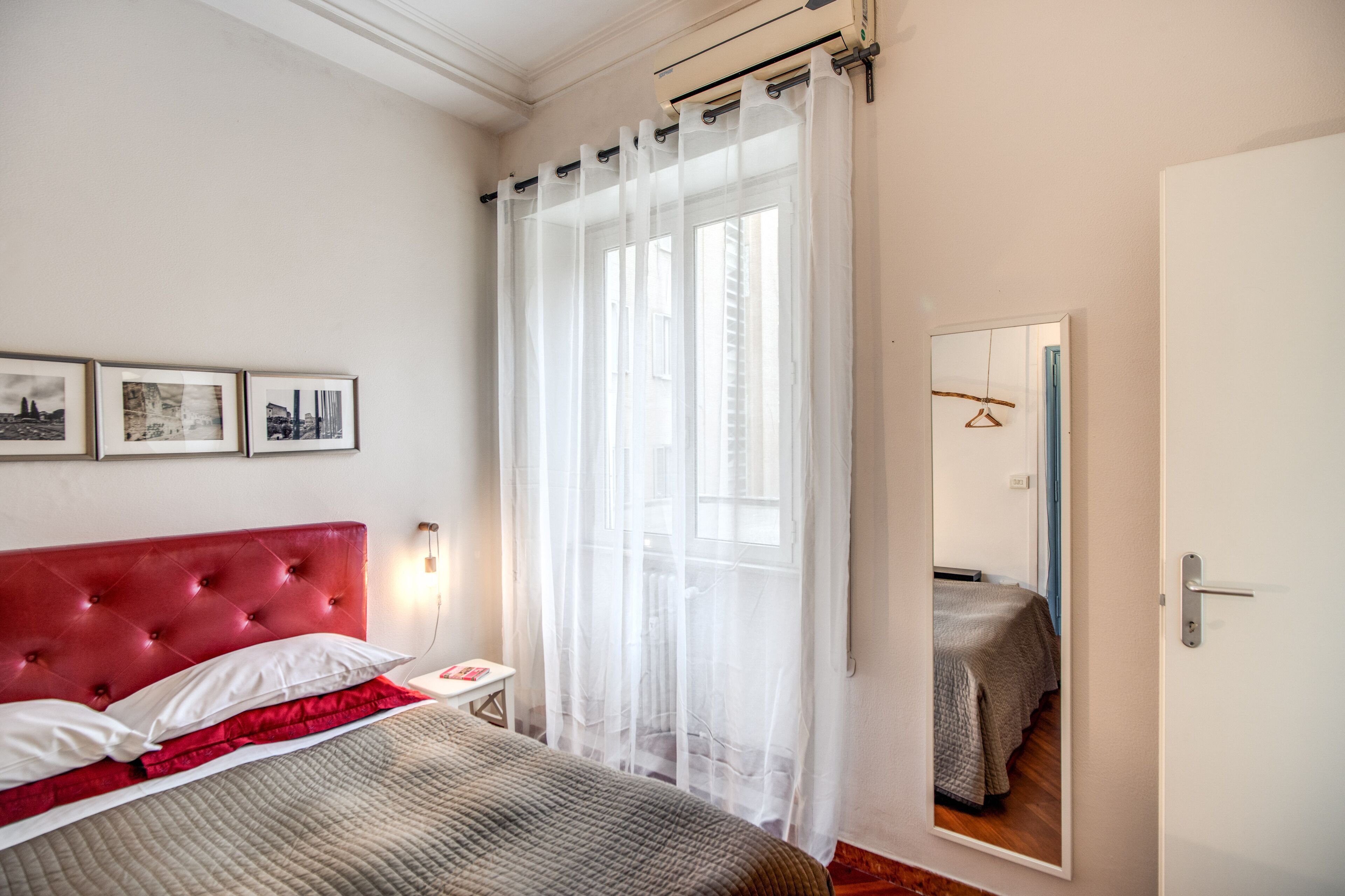 A bedroom with a red headboard, a bed with a grey quilt, a bedside table with a lamp, a window with sheer curtains, and a mirror on the wall.