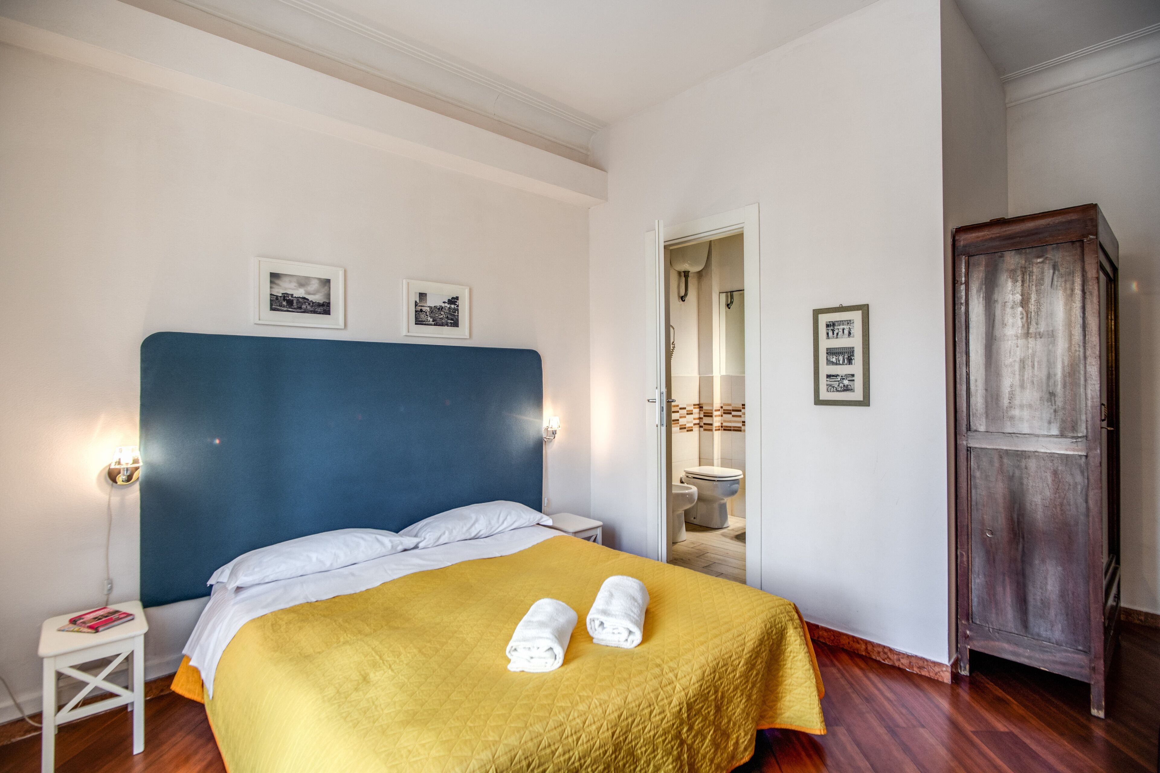 A hotel room with a large bed, a wooden wardrobe, and a small bedside table.