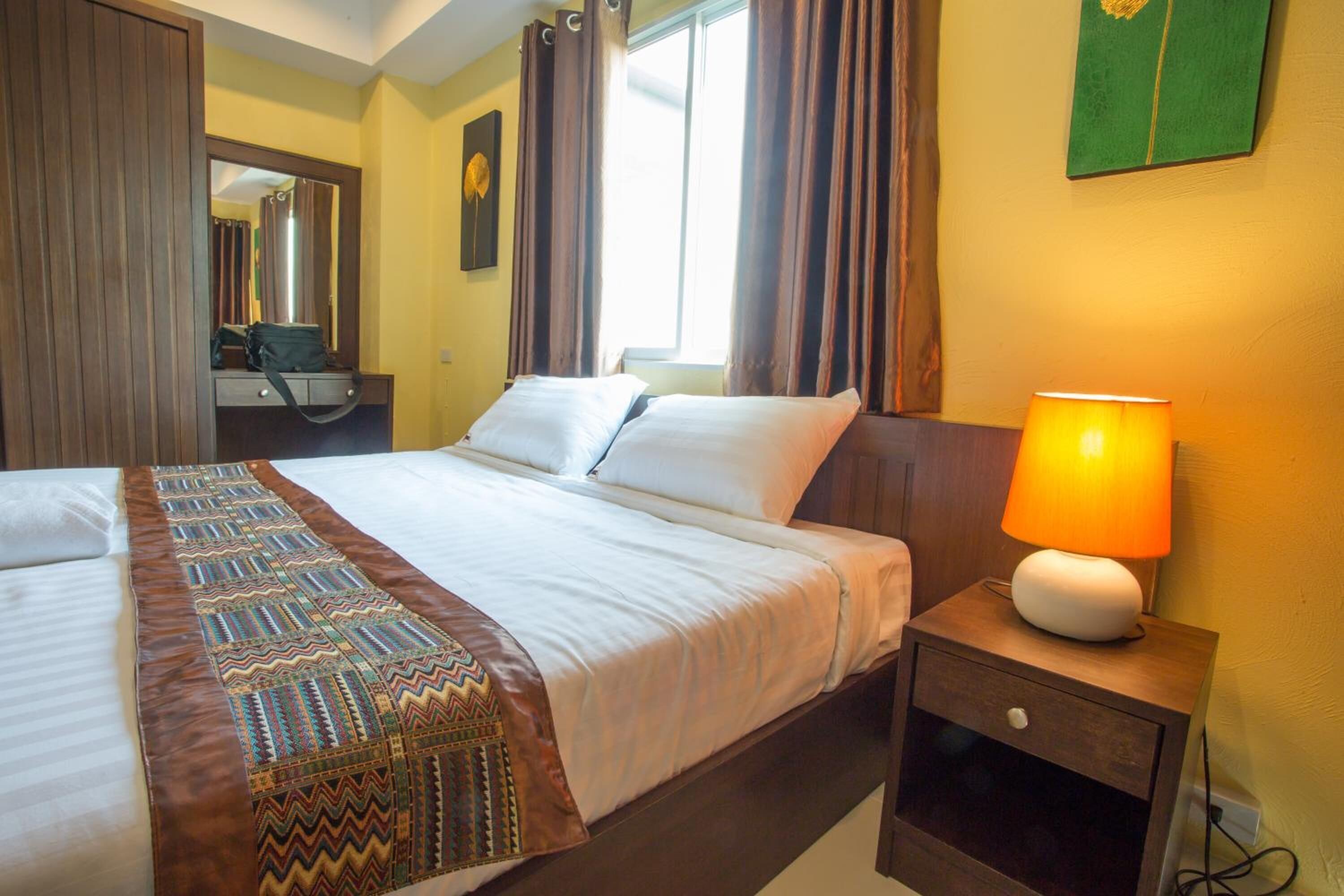 Une chambre d’hôtel comprenant un lit, une table de chevet avec une lampe, une commode, un miroir et une fenêtre avec des rideaux.