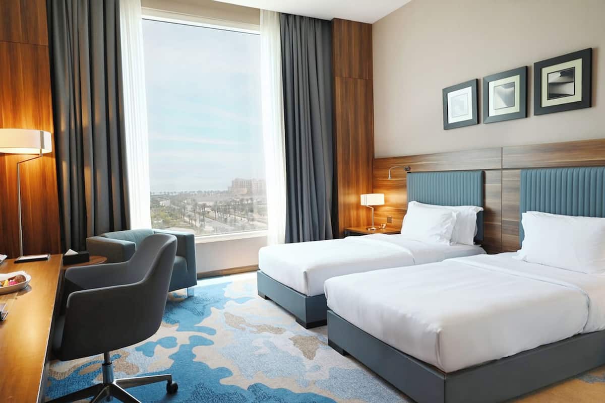 A hotel room with two beds, a desk with a chair, a large window with curtains, and framed pictures on the wall.