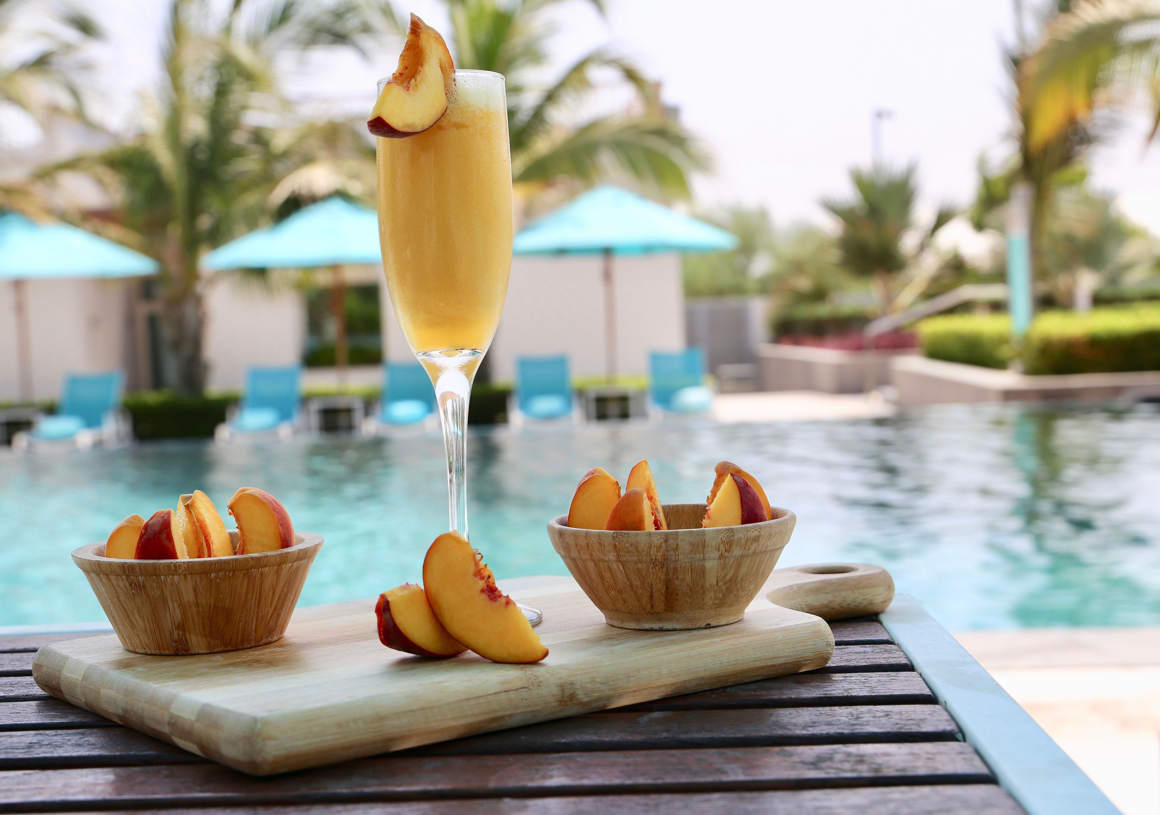 A poolside table with a glass of a peach-based cocktail, a wooden bowl with peach slices, and a wooden cutting board.