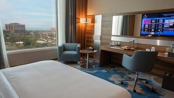 A hotel room with a bed, two armchairs, a desk with a computer, a TV, and a view of the city.