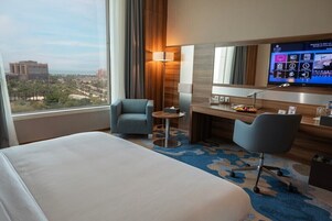 A hotel room with a bed, two armchairs, a desk with a computer, a TV, and a view of the city.