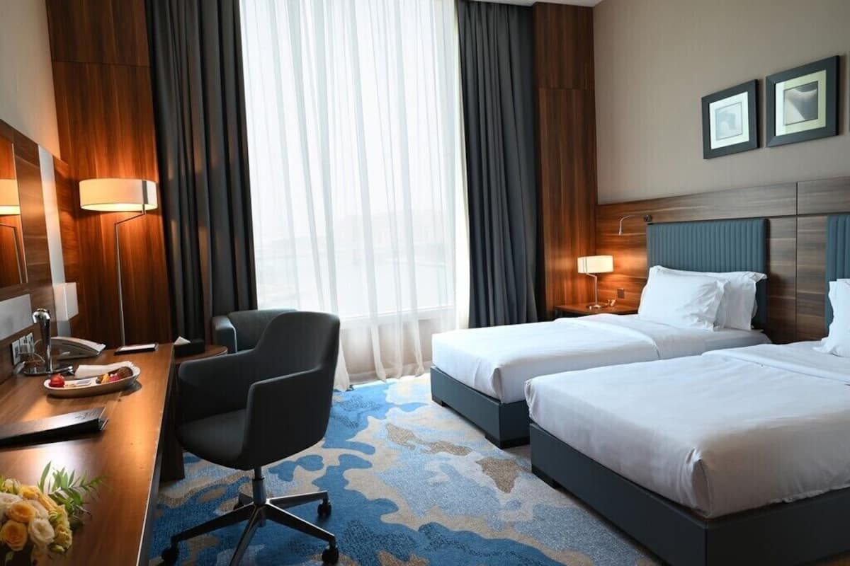 A hotel room with two beds, a desk with a chair, a lamp, and a large window with curtains.