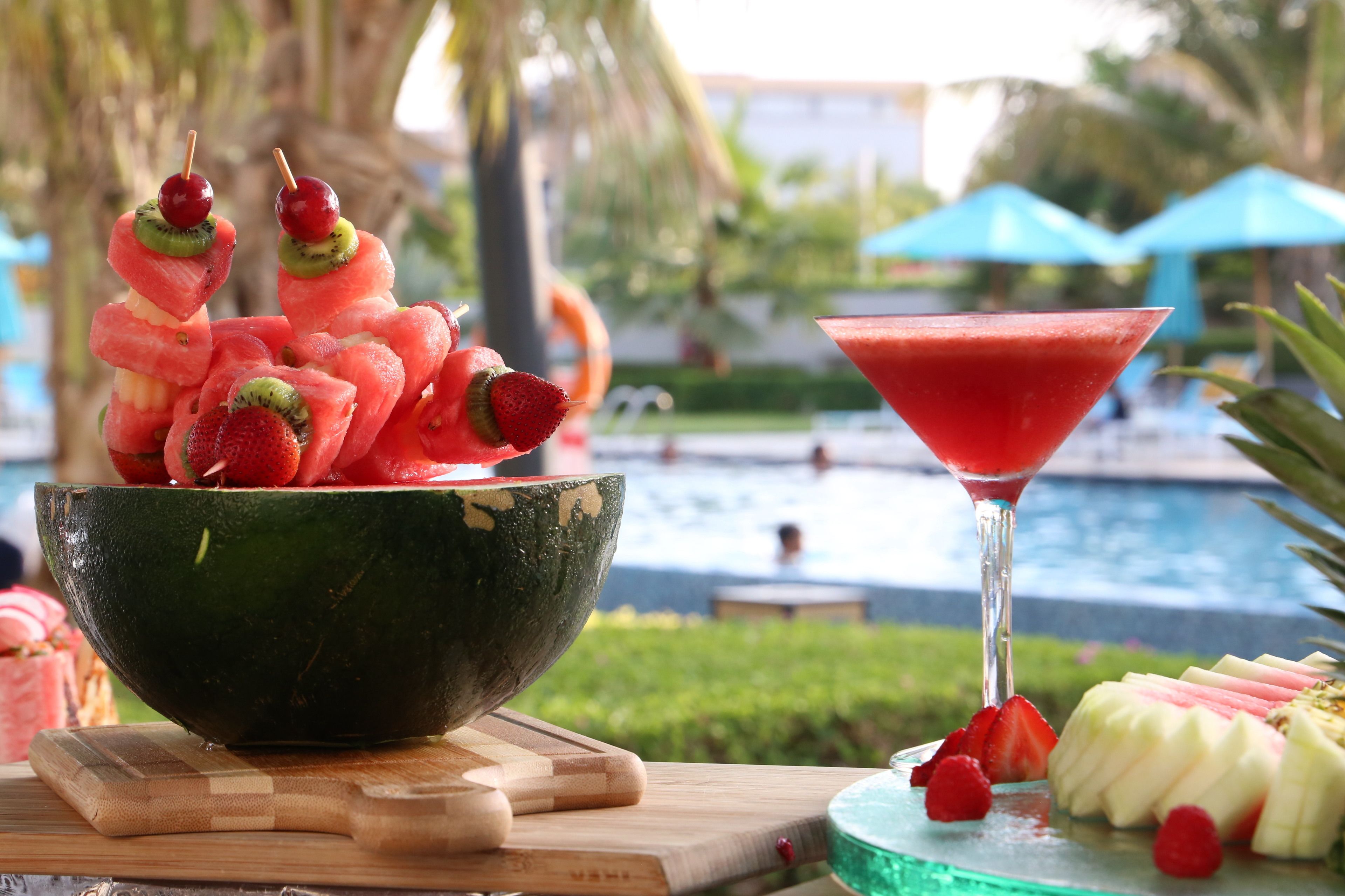 A bowl of fruit skewers and a martini glass on a wooden table with a pool view in the background.