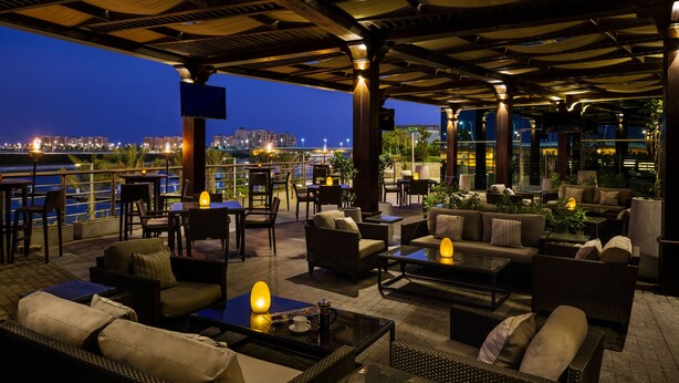 An outdoor seating area with tables and chairs, a TV screen, and a view of the city at night. - Bay La Sun Hotel & Marina (Rabigh)