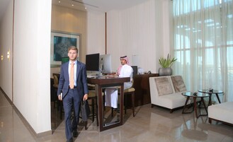 A man in a suit walking through a hotel lobby with a desk, computer, and a sofa.
