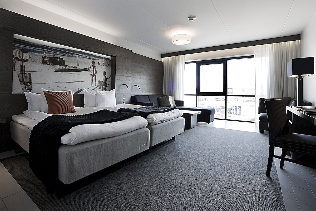 A modern hotel room with a large bed, a desk, and a view of the city through large windows.