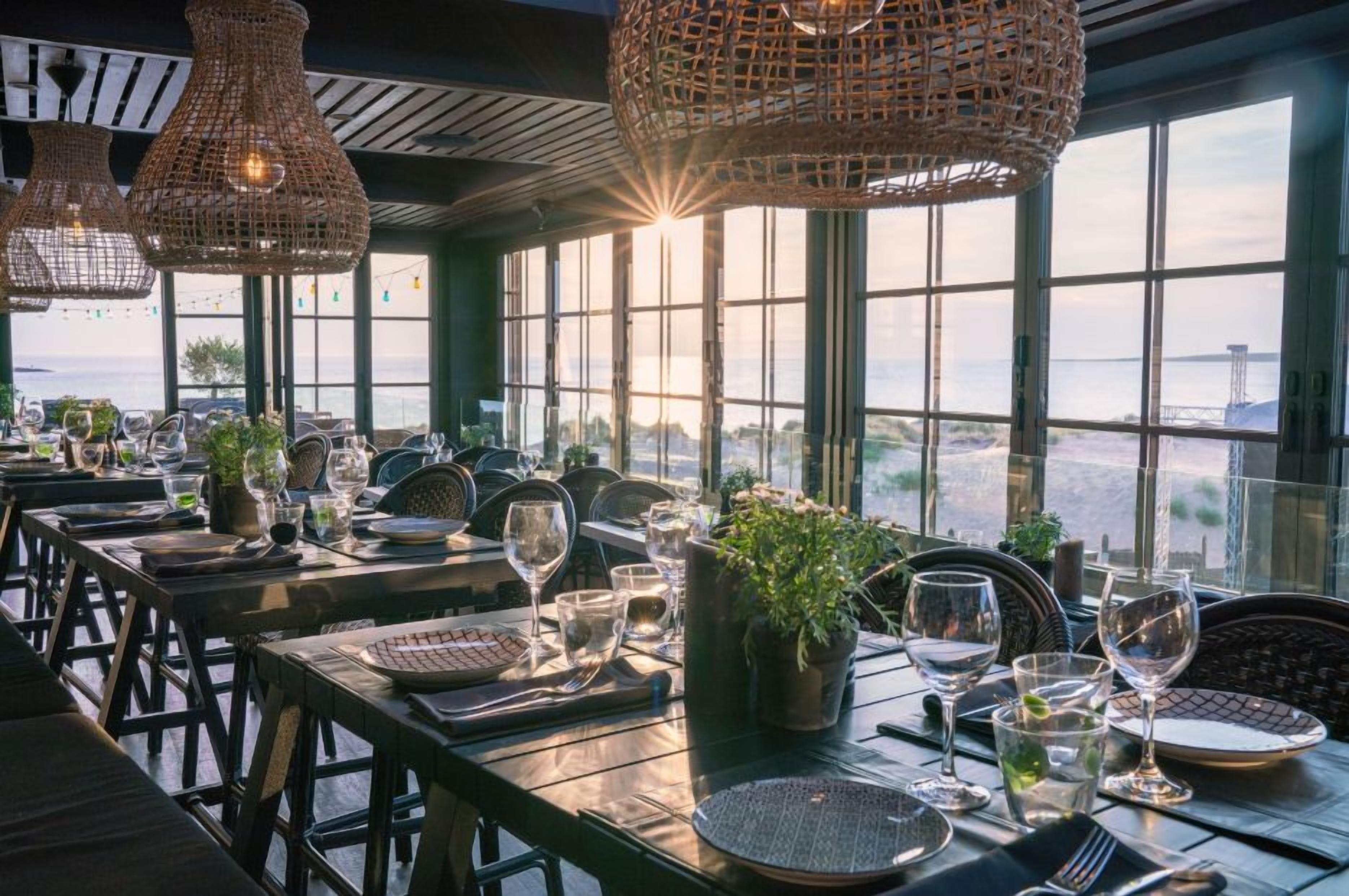 A restaurant with tables set for dining, large windows, and hanging wicker lamps.