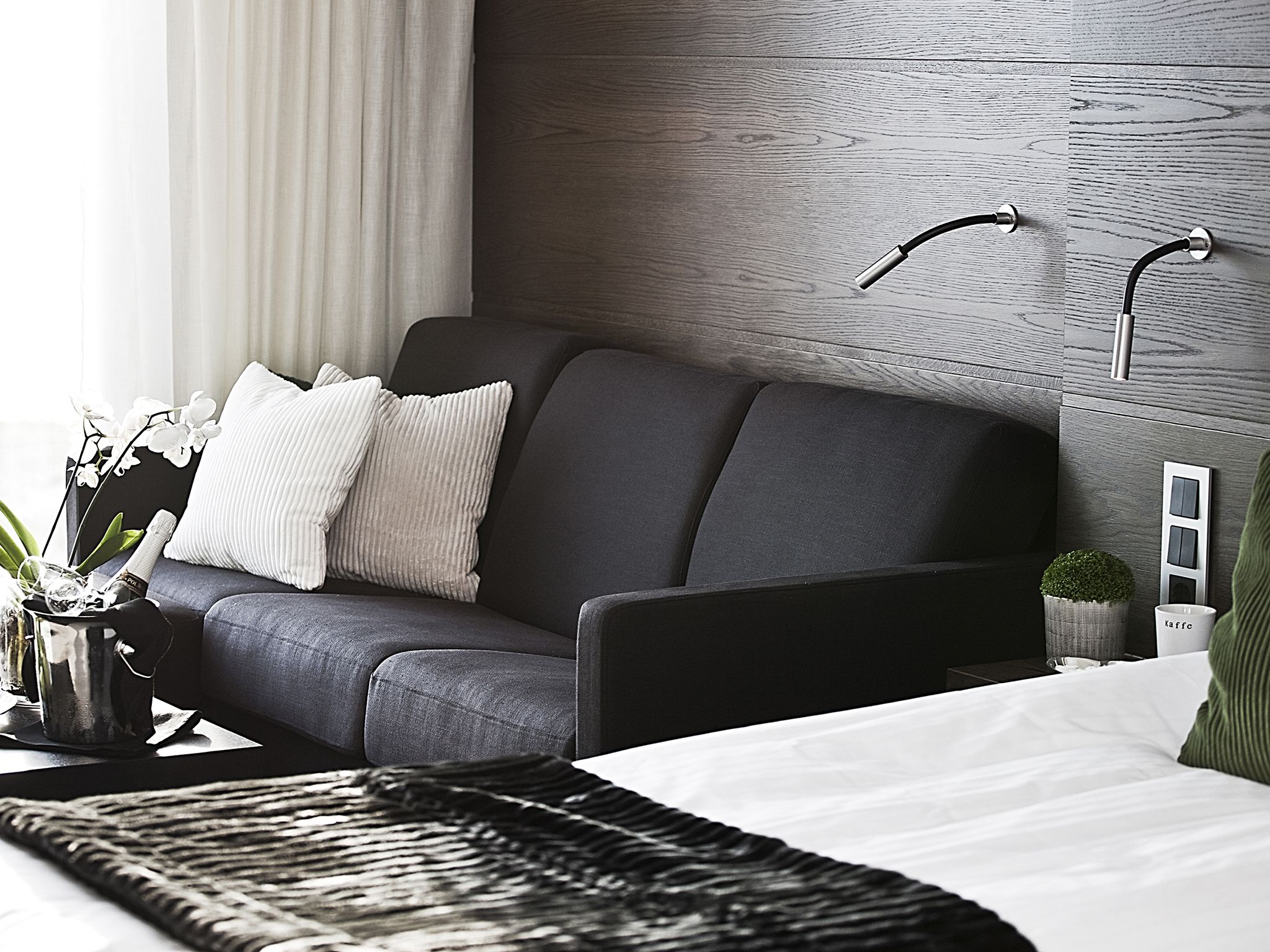 Una habitación de hotel moderna con un sofá gris, una cama con ropa de cama blanca, un cabecero de madera y una pequeña planta en maceta.