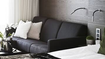 Una habitación de hotel moderna con un sofá gris, una cama con ropa de cama blanca, un cabecero de madera y una pequeña planta en maceta.
