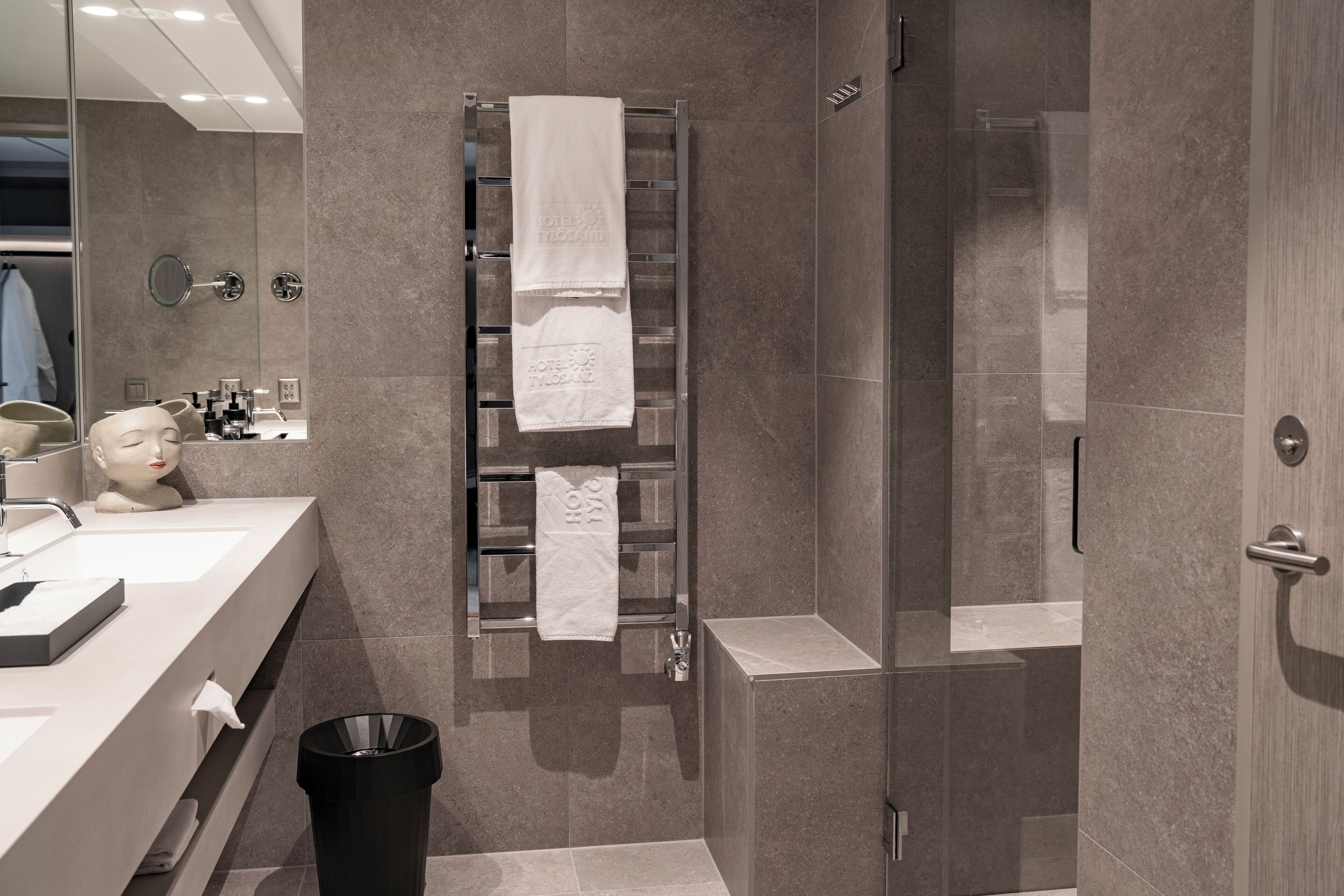 Un baño moderno con cabina de ducha de vidrio, un perchero con dos toallas, un tocador con espejo y un cubo de basura.