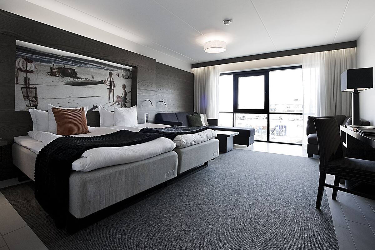 A modern hotel room with a large bed, a desk, and a view of the city through large windows.