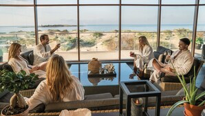 A group of people in a room with large windows overlooking a beach, sitting on sofas and chairs, with a table in the center. - Hotel Tylösand (Halmstad)