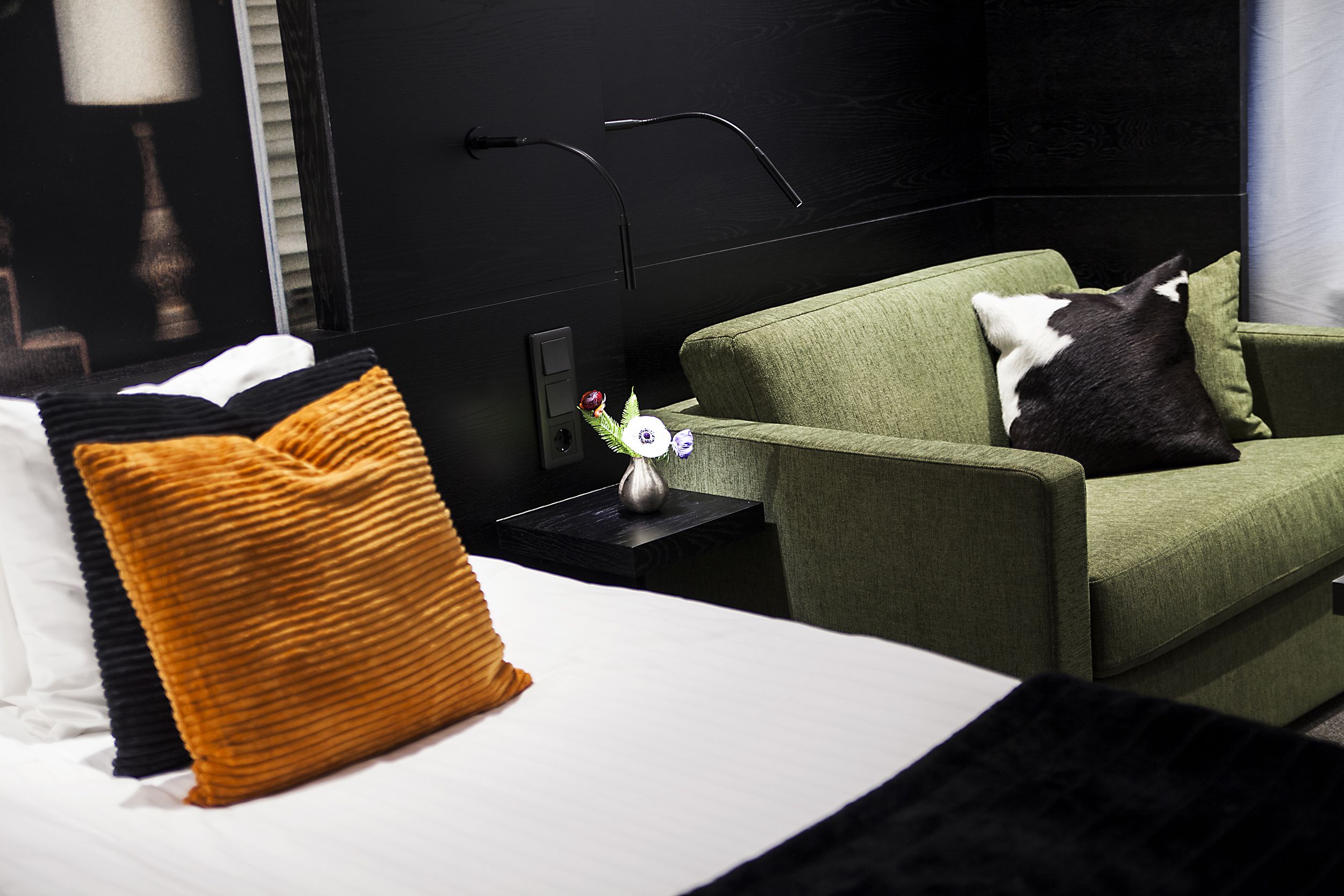 Ett hotellrum med en säng, en grön fåtölj, en svartvit katt och ett litet bord med en vas.