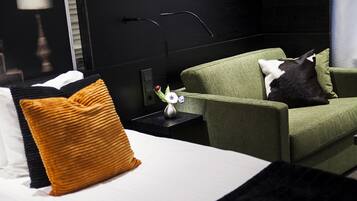 Una habitación de hotel con una cama, un sillón verde, un gato blanco y negro, y una pequeña mesa con un florero.