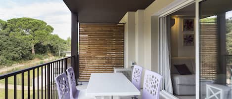 A balcony with a white table and purple chairs, a sliding glass door leading to an indoor seating area, and a view of greenery.