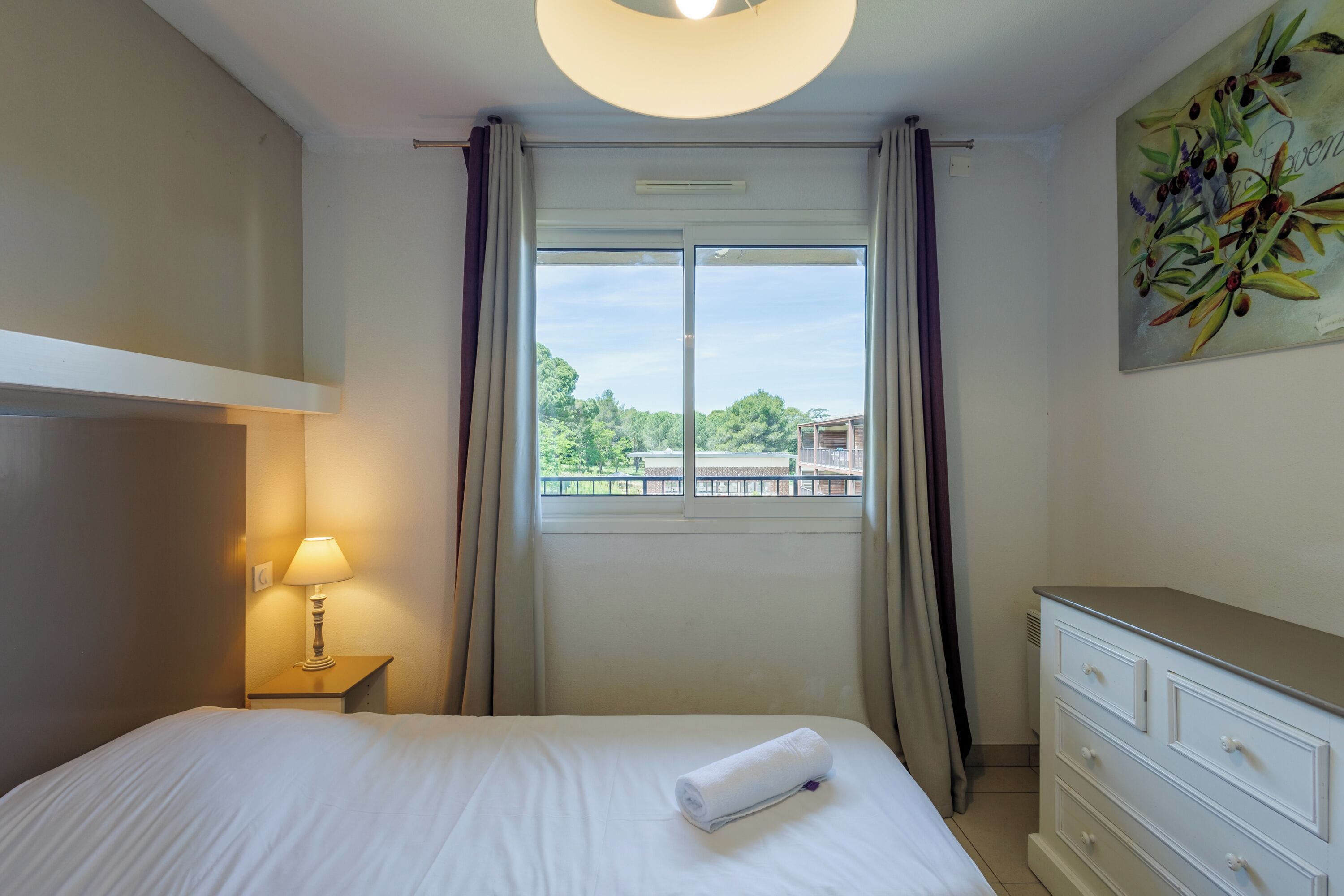 A hotel room with a bed, a nightstand with a lamp, a dresser, and a window with curtains.