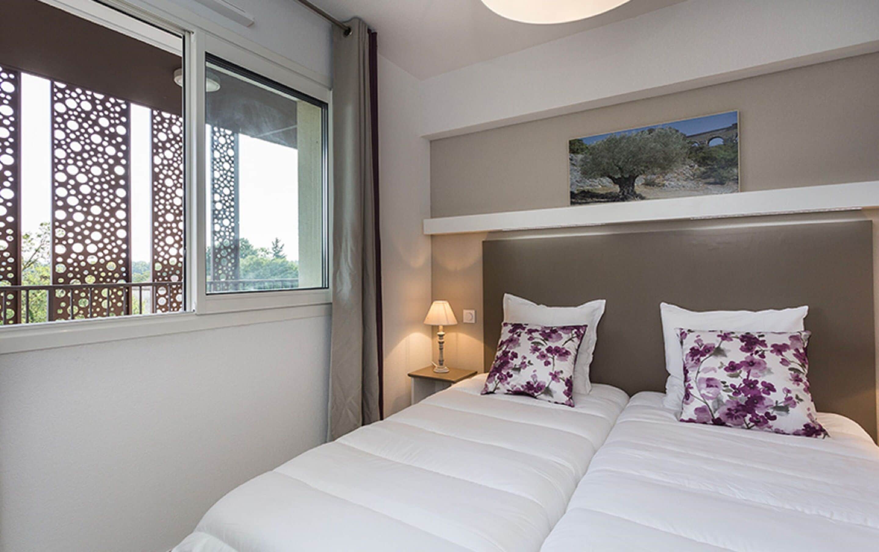 Habitación de hotel con cama, mesita de noche con lámpara, ventana con vista a árboles y un cuadro en la pared.