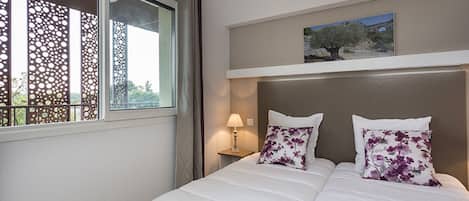 Una habitació d'hotel amb un llit, una taula de nit amb una làmpada, una finestra amb vistes als arbres i un quadre a la paret.