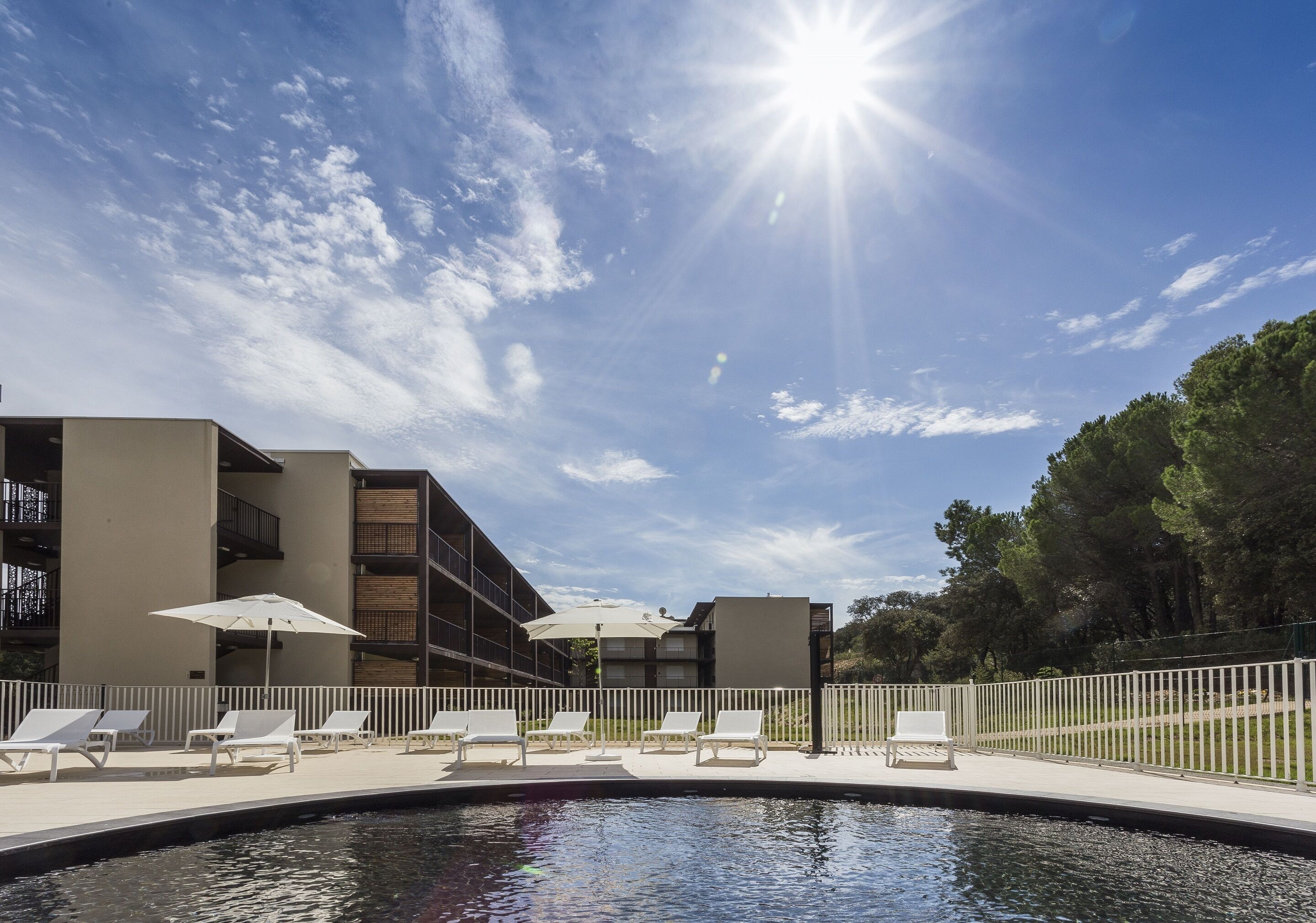 Zona de piscina con tumbonas y sombrillas, rodeada de una valla y edificios con balcones.