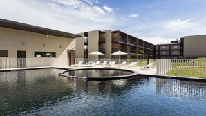 Piscina com espreguiçadeiras e guarda-sóis, cercada por um edifício com várias varandas.