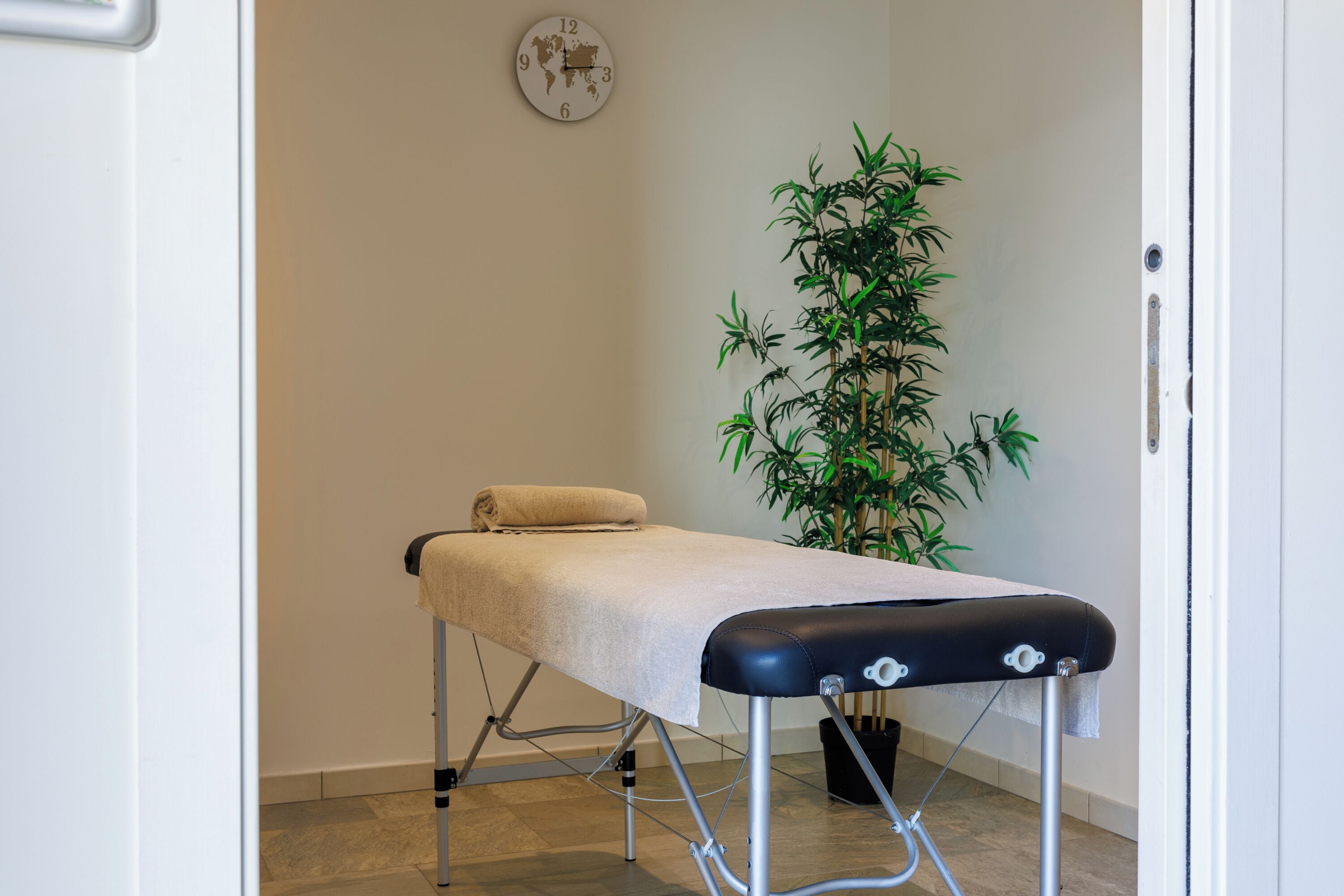 A massage table with a towel, a potted plant, and a world map clock on the wall.