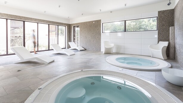 Uma área moderna de piscina interna com espreguiçadeiras, uma banheira de hidromassagem e janelas amplas com vista para o exterior.
