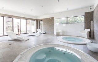 Uma área moderna de piscina interna com espreguiçadeiras, uma banheira de hidromassagem e janelas amplas com vista para o exterior.