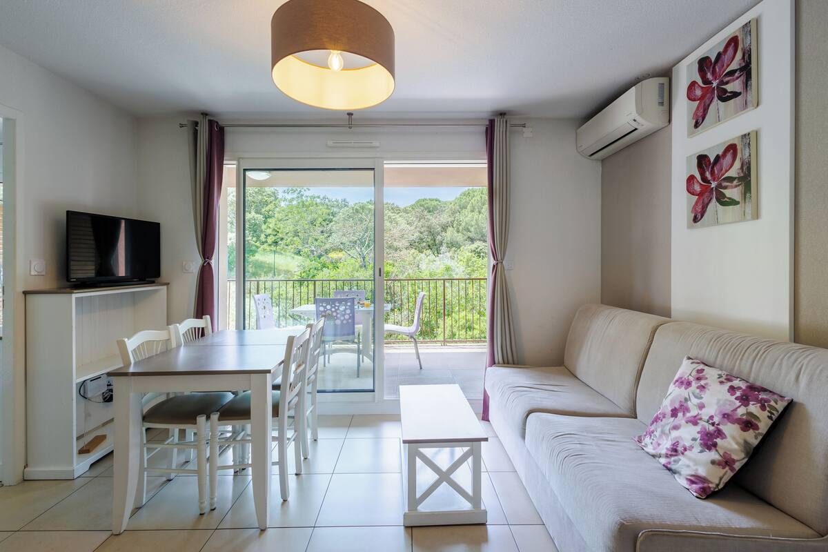 A modern living room with a sofa, a dining table, and chairs. There is a TV on a cabinet, and the room has a balcony with a view of greenery.