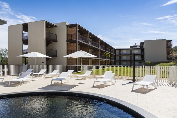 A poolside area with lounge chairs and umbrellas, a modern building with balconies, and a clear sky. - Residhotel – Résidence Pont du Gard (Remoulins)