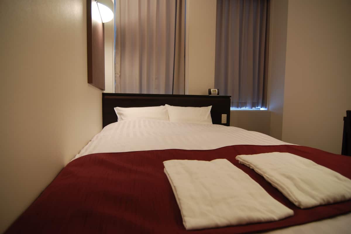 Cama bem arrumada com colcha vermelha e duas toalhas brancas, uma mesinha de cabeceira e uma janela com cortinas.