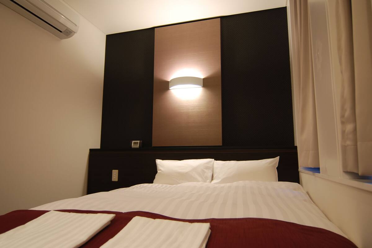 Quarto de hotel com cama, luminária na parede e janela com cortinas.