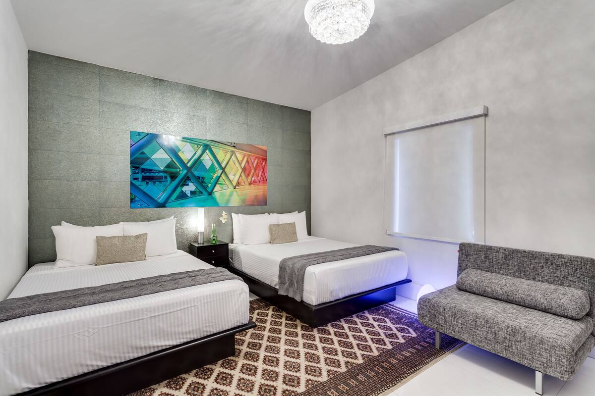 Una habitación de hotel moderna con dos camas, un sofá gris, un tapete estampado y una llamativa obra de arte geométrica en la pared.