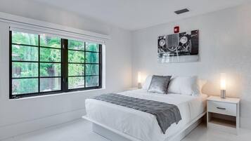 A modern bedroom with a large window, a bed with white bedding, a nightstand, and a wall-mounted clock.