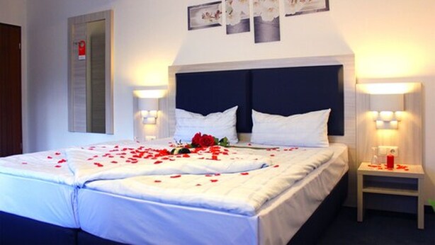 A neatly made bed with white linens and red rose petals, a dark headboard, and wall-mounted picture frames. - Hotel Altenwerder Hof (Hamburg)