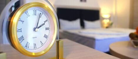 A clock with Roman numerals on a stand in a room with a bed and a wall-mounted light.