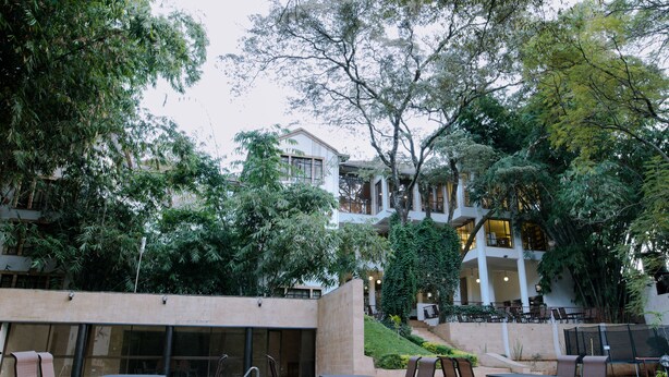 An outdoor patio area with tables and chairs, surrounded by lush greenery and a building with multiple floors and large windows. - West Wood Hotel (Nairobi)
