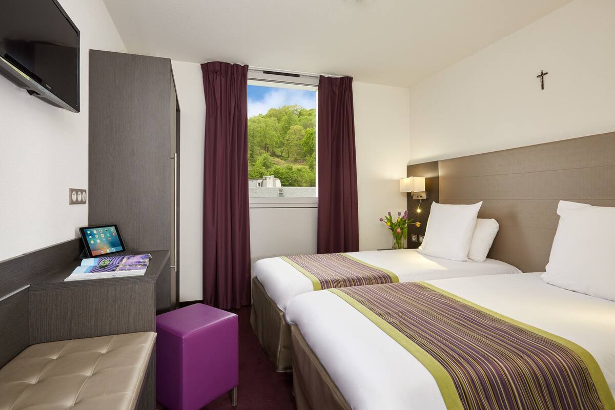 A hotel room with two beds, a desk with a tablet, a purple chair, and a window with curtains.